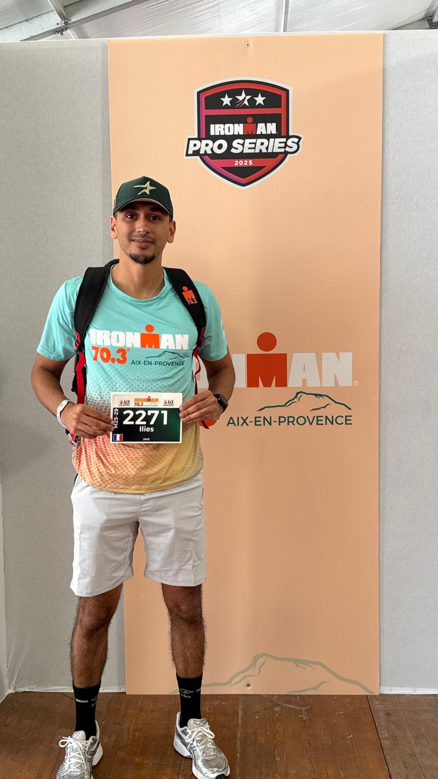 Illiès Chebil devant le panneau IRONMAN 70.3 Aix-en-Provence, son dossard à la main.
